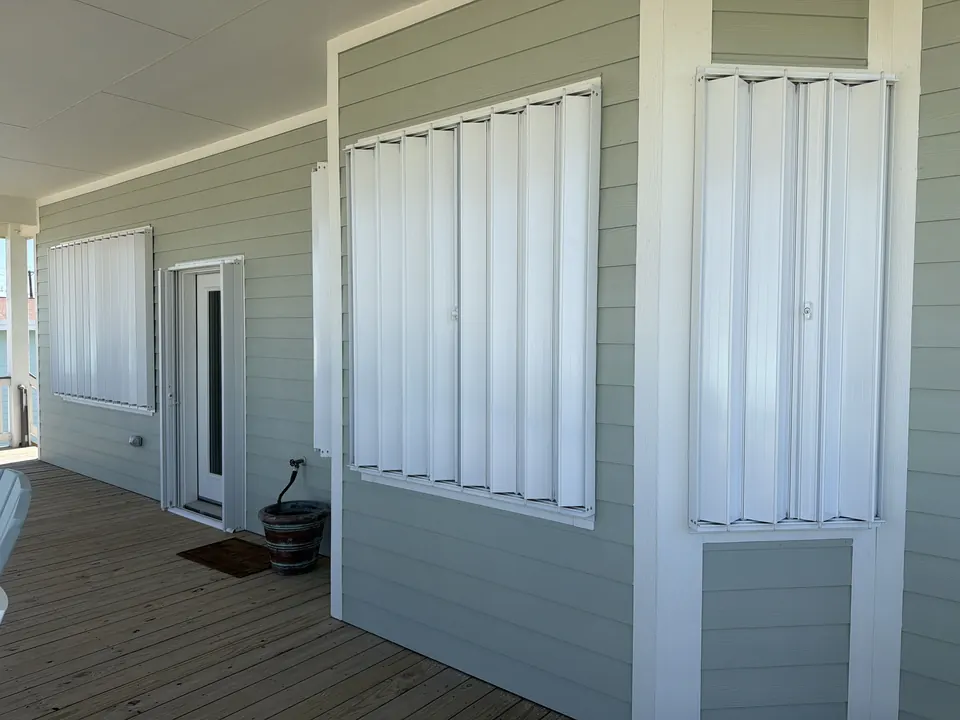 White accordion storm shutters closed over two windows on a sage-green covered porch