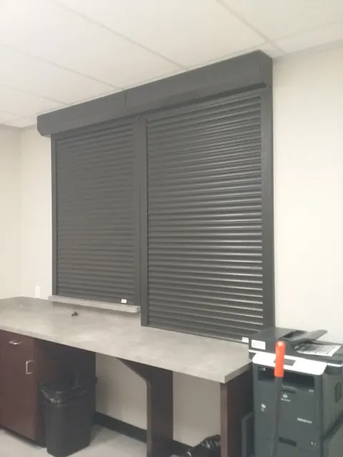 Two bronze roll-down security shutters closed over a reception counter window inside a clinic in Angleton
