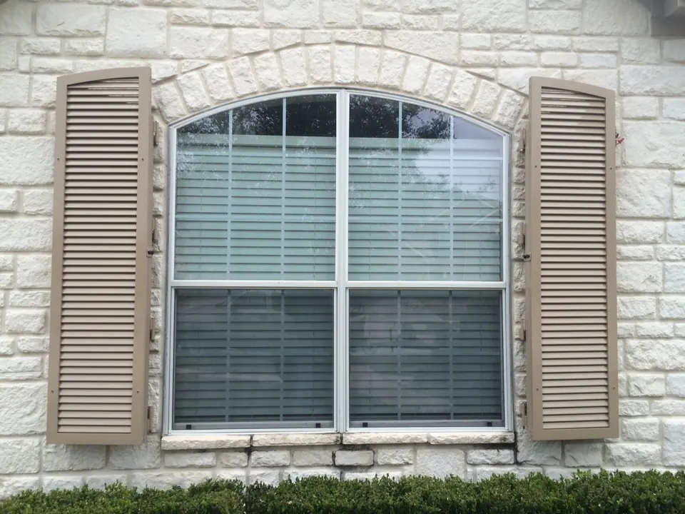 Tan louvered colonial shutters beside an arched window on a white stone home in Friendswood