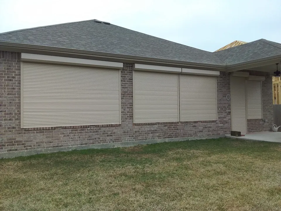 Tan roll-down shutters closed over the windows of a brick home in Seabrook