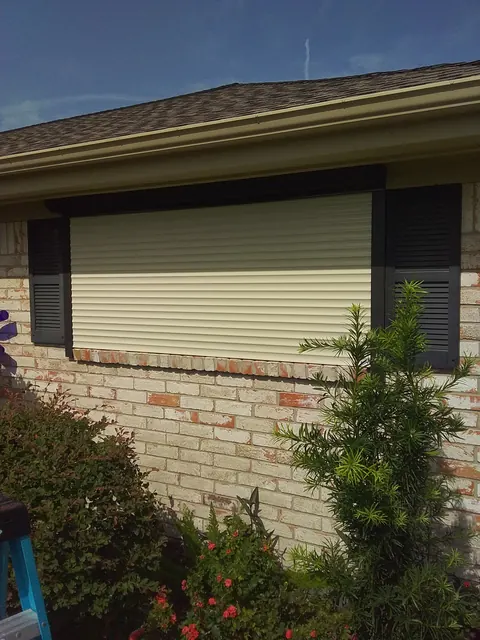 A tan roll-down shutter closed over a window on a brick home in Texas City