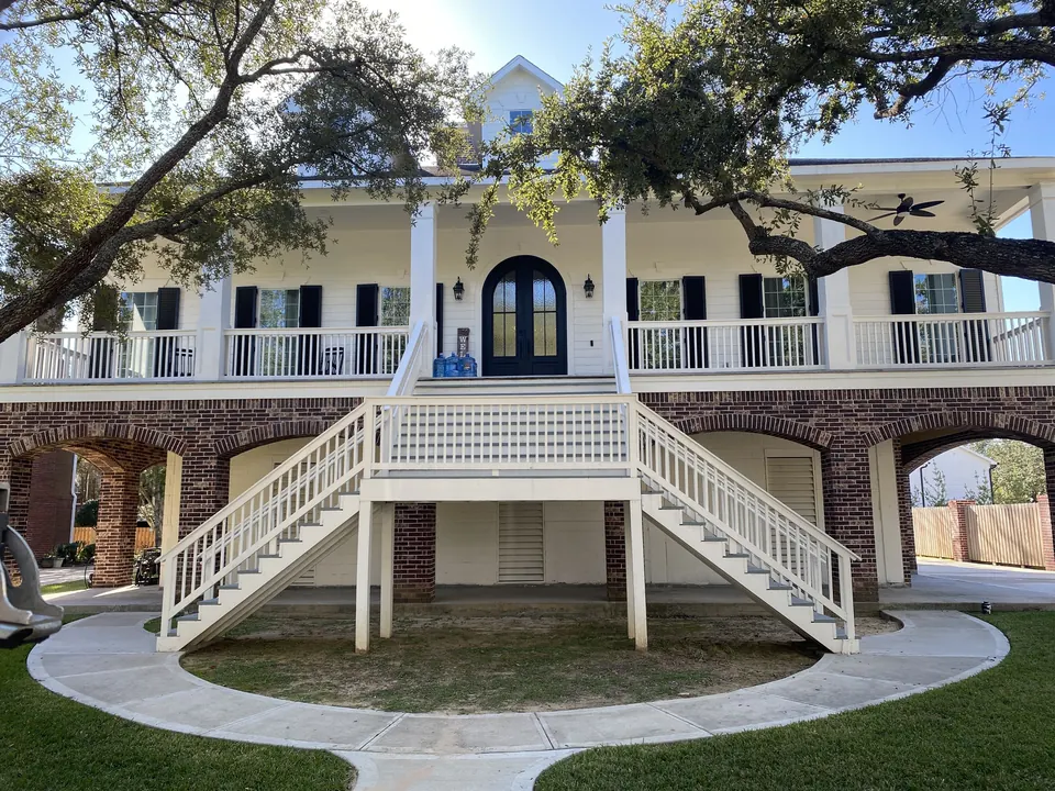 White raised plantation-style house with black louvered colonial shutters behind live oak branches