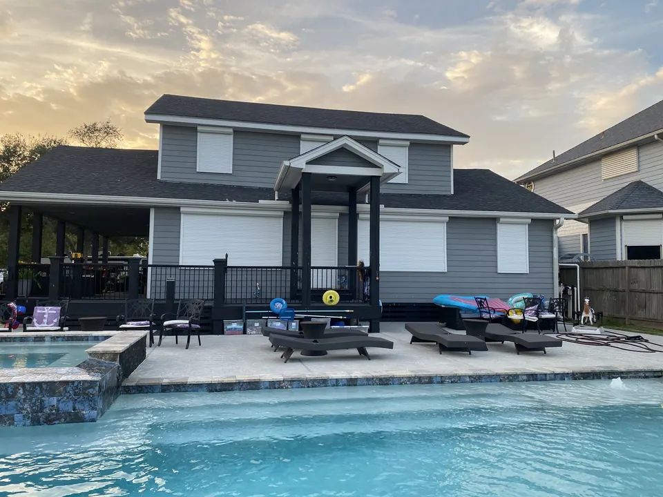White roll-down shutters closed across the back of a gray two-story home behind a pool